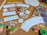 A collection of board game components and crafting materials are spread out on a cork board surface. There are playing cards, dice in various colors and sizes, wooden letter tiles spelling 'BOARDGAMES', 'DRAW', and 'WIN', colored buttons, a variety of markers and colored pencils, scissors, glue stick, and circular cardboard cutouts. The setting is vibrant and full of an assortment of gaming and crafting items.
