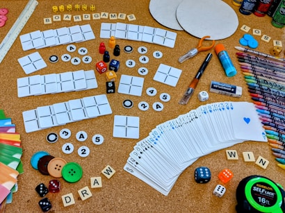 A collection of board game components and crafting materials are spread out on a cork board surface. There are playing cards, dice in various colors and sizes, wooden letter tiles spelling 'BOARDGAMES', 'DRAW', and 'WIN', colored buttons, a variety of markers and colored pencils, scissors, glue stick, and circular cardboard cutouts. The setting is vibrant and full of an assortment of gaming and crafting items.