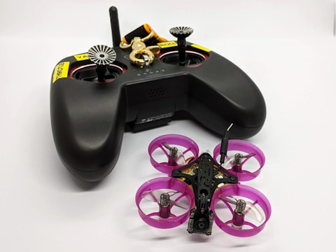 A black remote control with dual joysticks is positioned beside a small quadcopter drone. The drone features vivid pink propeller guards and has a compact, lightweight design, indicating it might be designed for indoor or recreational use. A keychain with a golden key and a black and orange lanyard is attached to the remote.