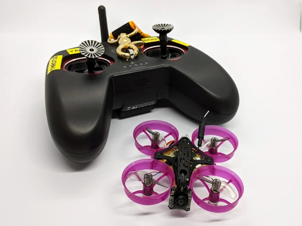 A black remote control with dual joysticks is positioned beside a small quadcopter drone. The drone features vivid pink propeller guards and has a compact, lightweight design, indicating it might be designed for indoor or recreational use. A keychain with a golden key and a black and orange lanyard is attached to the remote.