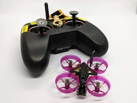 A black remote control with dual joysticks is positioned beside a small quadcopter drone. The drone features vivid pink propeller guards and has a compact, lightweight design, indicating it might be designed for indoor or recreational use. A keychain with a golden key and a black and orange lanyard is attached to the remote.