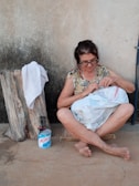 An older woman with glasses is sitting on a cushion on a cement floor, focused on sewing a white textile with a red stripe. Her legs are crossed, and she is barefoot. Next to her is a tree stump with a white towel draped over it and a blue and white canister of Molico. The background consists of a plain textured wall.