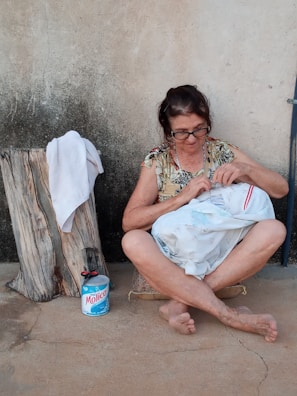 An older woman with glasses is sitting on a cushion on a cement floor, focused on sewing a white textile with a red stripe. Her legs are crossed, and she is barefoot. Next to her is a tree stump with a white towel draped over it and a blue and white canister of Molico. The background consists of a plain textured wall.
