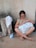 An older woman with glasses is sitting on a cushion on a cement floor, focused on sewing a white textile with a red stripe. Her legs are crossed, and she is barefoot. Next to her is a tree stump with a white towel draped over it and a blue and white canister of Molico. The background consists of a plain textured wall.