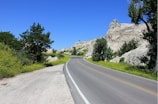 A scenic road curves through a landscape featuring rocky formations and lush greenery. Trees and bushes line the roadside, and the sky is clear with a vibrant blue hue.