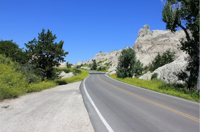 A scenic road curves through a landscape featuring rocky formations and lush greenery. Trees and bushes line the roadside, and the sky is clear with a vibrant blue hue.