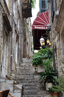A narrow stone staircase leads upward between old stone buildings. A red canopy with the words 'RAGUSA 2' is prominently displayed on the right. Green plants in pots line the staircase, adding a touch of color to the scene. The walls are adorned with rustic lanterns and various wires, evoking an aged and charming atmosphere.