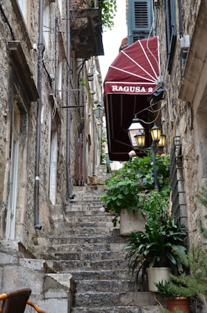 A narrow stone staircase leads upward between old stone buildings. A red canopy with the words 'RAGUSA 2' is prominently displayed on the right. Green plants in pots line the staircase, adding a touch of color to the scene. The walls are adorned with rustic lanterns and various wires, evoking an aged and charming atmosphere.