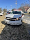 A white Chevrolet SUV with red and white emergency lights is parked on a grassy area near a road. Power lines and utility poles are visible behind it, along with several buildings and trees in the distance. The vehicle has a license plate and displays a sign indicating it is a fire department vehicle.