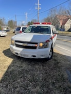 A white Chevrolet SUV with red and white emergency lights is parked on a grassy area near a road. Power lines and utility poles are visible behind it, along with several buildings and trees in the distance. The vehicle has a license plate and displays a sign indicating it is a fire department vehicle.