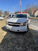 A white Chevrolet SUV with red and white emergency lights is parked on a grassy area near a road. Power lines and utility poles are visible behind it, along with several buildings and trees in the distance. The vehicle has a license plate and displays a sign indicating it is a fire department vehicle.