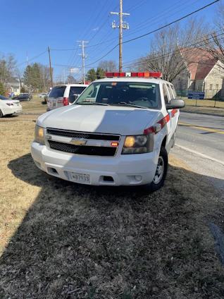 A white Chevrolet SUV with red and white emergency lights is parked on a grassy area near a road. Power lines and utility poles are visible behind it, along with several buildings and trees in the distance. The vehicle has a license plate and displays a sign indicating it is a fire department vehicle.