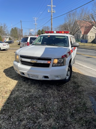 A white Chevrolet SUV with red and white emergency lights is parked on a grassy area near a road. Power lines and utility poles are visible behind it, along with several buildings and trees in the distance. The vehicle has a license plate and displays a sign indicating it is a fire department vehicle.