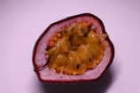 A halved passion fruit showing its vibrant yellow and orange pulpy interior with numerous small black seeds. The exterior rind is a deep reddish-purple color, and the surface of the fruit has a glossy texture.