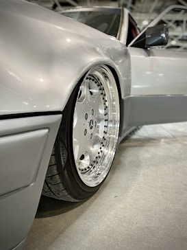 A close-up view of a polished silver car wheel with a chrome rim, featuring detailed lug nuts. The car body is a sleek metallic silver, conveying a sense of luxury and precision engineering. The image captures a modern vehicle inside a well-lit indoor setting.