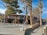 Outdoor market scene with a stall displaying an array of colorful clothing items such as shirts and dresses. Trees provide shade over the market area, while a sign directs towards restrooms. Visitors browse through the merchandise under a clear blue sky.