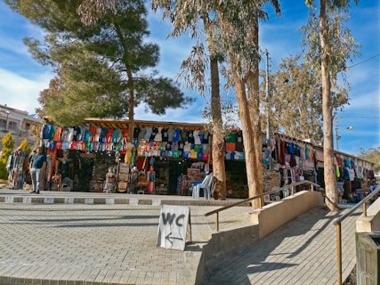 Outdoor market scene with a stall displaying an array of colorful clothing items such as shirts and dresses. Trees provide shade over the market area, while a sign directs towards restrooms. Visitors browse through the merchandise under a clear blue sky.
