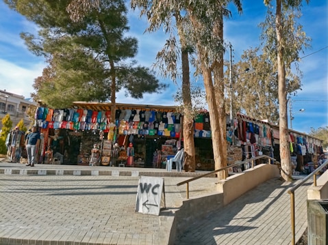 Outdoor market scene with a stall displaying an array of colorful clothing items such as shirts and dresses. Trees provide shade over the market area, while a sign directs towards restrooms. Visitors browse through the merchandise under a clear blue sky.