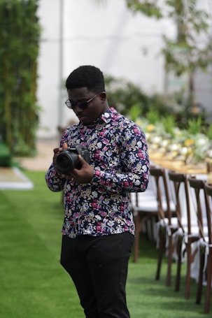 A man with a patterned floral shirt holds a camera while standing on a grass lawn. In the background, there is a long table set up with chairs and decorative plants, suggesting a festive or formal outdoor event.