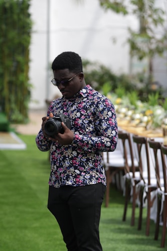 A man with a patterned floral shirt holds a camera while standing on a grass lawn. In the background, there is a long table set up with chairs and decorative plants, suggesting a festive or formal outdoor event.