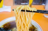A close-up view of a bowl of ramen noodles being lifted with black chopsticks. The soup broth appears dark, and some seaweed is visible on the side of the bowl, set against an orange background.