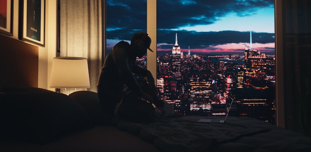 a man sitting on a bed looking out a window at the city