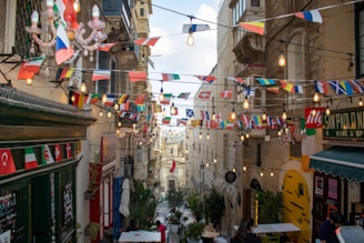 A narrow street in a European setting is adorned with multiple colorful international flags hanging across, adding a festive atmosphere. Warm light bulbs are strung overhead, creating a cozy ambiance. The architecture features rustic, old stone buildings with balconies, and people can be seen walking or sitting outside at cafes or restaurants along the street.