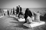 Several individuals are gathered in an outdoor setting, with many of them seated on the ground alongside bags and containers of supplies, likely food aid. The people are dressed in cultural attire, and there is a visible stone wall in the background dividing the scene.