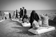 Several individuals are gathered in an outdoor setting, with many of them seated on the ground alongside bags and containers of supplies, likely food aid. The people are dressed in cultural attire, and there is a visible stone wall in the background dividing the scene.