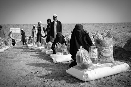 Several individuals are gathered in an outdoor setting, with many of them seated on the ground alongside bags and containers of supplies, likely food aid. The people are dressed in cultural attire, and there is a visible stone wall in the background dividing the scene.