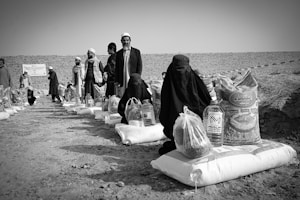 Several individuals are gathered in an outdoor setting, with many of them seated on the ground alongside bags and containers of supplies, likely food aid. The people are dressed in cultural attire, and there is a visible stone wall in the background dividing the scene.