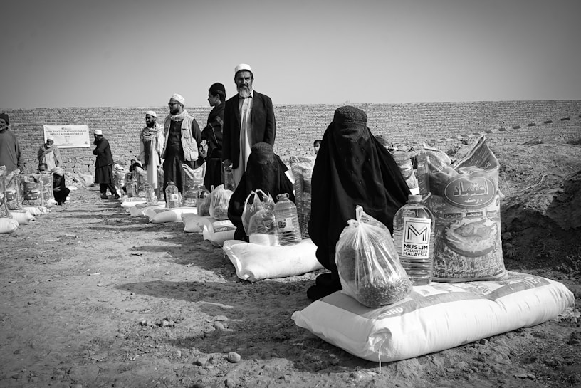 Several individuals are gathered in an outdoor setting, with many of them seated on the ground alongside bags and containers of supplies, likely food aid. The people are dressed in cultural attire, and there is a visible stone wall in the background dividing the scene.