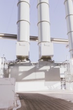 Three large industrial structures dominate the scene, resembling tall, cylindrical silos or chimneys. They are positioned vertically and appear to be part of an extensive industrial setup, possibly related to power generation or distribution. The environment features a clear sky and well-lit conditions, suggesting it might be outdoors.