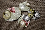Several crushed aluminum beverage cans lie on a textured carpet. The cans appear to be from different brands, with some displaying remnants of colorful labels and distinctive pull tabs. The background is made up of a brown and beige patterned carpet.