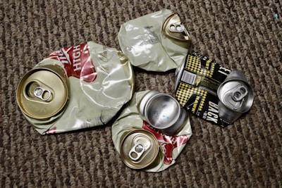 Several crushed aluminum beverage cans lie on a textured carpet. The cans appear to be from different brands, with some displaying remnants of colorful labels and distinctive pull tabs. The background is made up of a brown and beige patterned carpet.