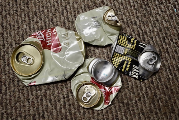 Several crushed aluminum beverage cans lie on a textured carpet. The cans appear to be from different brands, with some displaying remnants of colorful labels and distinctive pull tabs. The background is made up of a brown and beige patterned carpet.