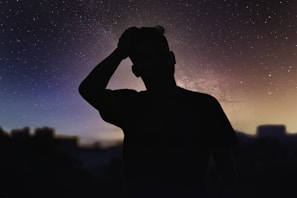 A silhouette of a person with a hand on their head stands against a starry night sky. The milky way is faintly visible, creating a dreamy and serene atmosphere. The person appears contemplative or relaxed.