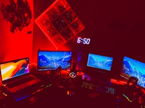 A dimly lit room featuring a setup with multiple computer screens displaying vibrant graphics. The area is illuminated by red LED lights, creating a futuristic and intense ambiance. There is a digital clock showing 6:50 mounted on the wall, and various tech accessories are visible on the desk. The red lighting dominates the scene, giving the space a high-tech, gaming environment feel.