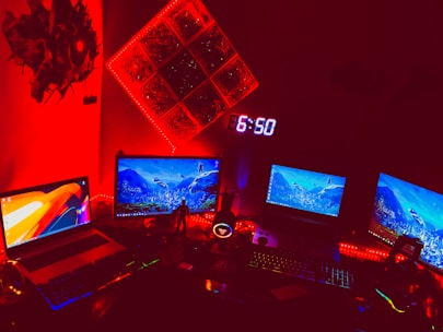 A dimly lit room featuring a setup with multiple computer screens displaying vibrant graphics. The area is illuminated by red LED lights, creating a futuristic and intense ambiance. There is a digital clock showing 6:50 mounted on the wall, and various tech accessories are visible on the desk. The red lighting dominates the scene, giving the space a high-tech, gaming environment feel.
