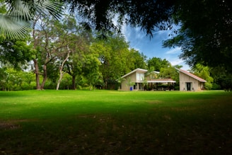 A spacious lawn stretches in front of a modern house with a unique architectural design, featuring two peaked roofs. The house is surrounded by lush greenery and large trees, under a partly cloudy blue sky.