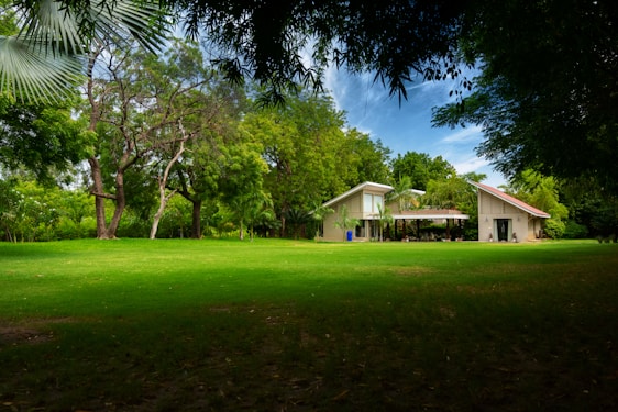 A spacious lawn stretches in front of a modern house with a unique architectural design, featuring two peaked roofs. The house is surrounded by lush greenery and large trees, under a partly cloudy blue sky.