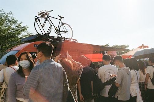 A group of people, some wearing masks, stand closely together around an orange vehicle with a bicycle mounted on the roof. The scene is outdoors, with the sun casting lens flare across the photo. There are trees in the background and some people holding umbrellas.