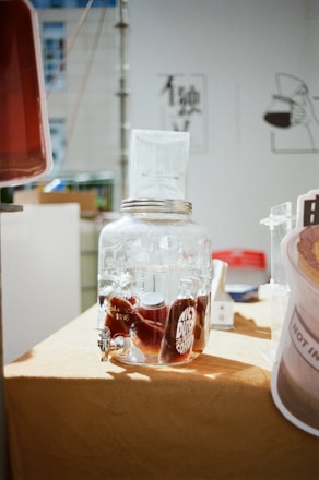 A glass dispenser filled with dark liquid, likely coffee, is placed on a light brown tablecloth. A paper filter is perched on top of the dispenser. In the background, there are blurred images of signage and other objects in a well-lit room.