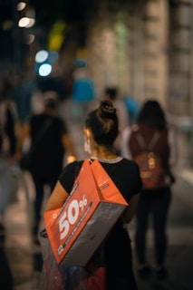 A person is walking down a dimly lit street carrying a large red shopping bag advertising a 50% discount. The street is bustling with other pedestrians, and the background features blurred lights and buildings, creating a sense of movement and activity.