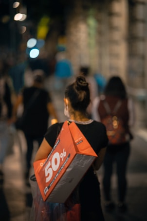 A person is walking down a dimly lit street carrying a large red shopping bag advertising a 50% discount. The street is bustling with other pedestrians, and the background features blurred lights and buildings, creating a sense of movement and activity.