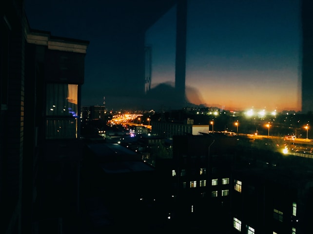 The image captures an urban landscape at dusk with city lights illuminating the streets. In the foreground, buildings are partially lit, while the horizon displays a gradient of deep blue to orange, suggesting sunset. Reflective surfaces and streetlights add warmth and contrast to the scene.