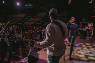 A lively performance captured with a musician playing a guitar and another person singing into a microphone on stage. The audience, visible in the background, is engaged and appears enthusiastic, with many raising their hands or wearing masks. Stage lights create a dramatic ambiance with patterns on the floor.