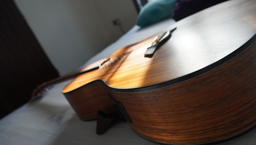 A close-up view of an acoustic guitar lying flat on a bed, capturing the side and body of the instrument. Soft natural light highlights the wooden texture and grain, adding warmth to the scene. The background is blurred, emphasizing the focus on the guitar.