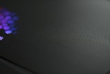 A close-up view of a perforated metal surface with a reflective texture. Subtle purple and blue light glimmers on one side, creating a dynamic contrast against the dark, matte finish of the surface.