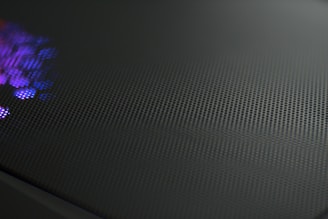 A close-up view of a perforated metal surface with a reflective texture. Subtle purple and blue light glimmers on one side, creating a dynamic contrast against the dark, matte finish of the surface.